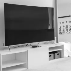 Duży telewizor, prawdopodobnie o wymiarach telewizora 75 cali, stoi na białej szafce RTV. W tle lustro z kalendarzem.