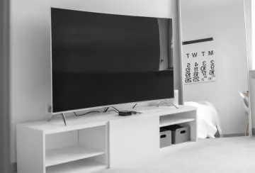 Duży telewizor, prawdopodobnie o wymiarach telewizora 75 cali, stoi na białej szafce RTV. W tle lustro z kalendarzem.